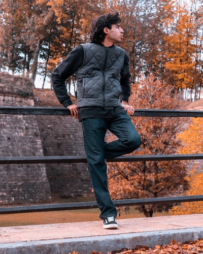 一个穿着黑色毛衣、灰色镀金衫和蓝色牛仔裤靠在栏杆上的男人